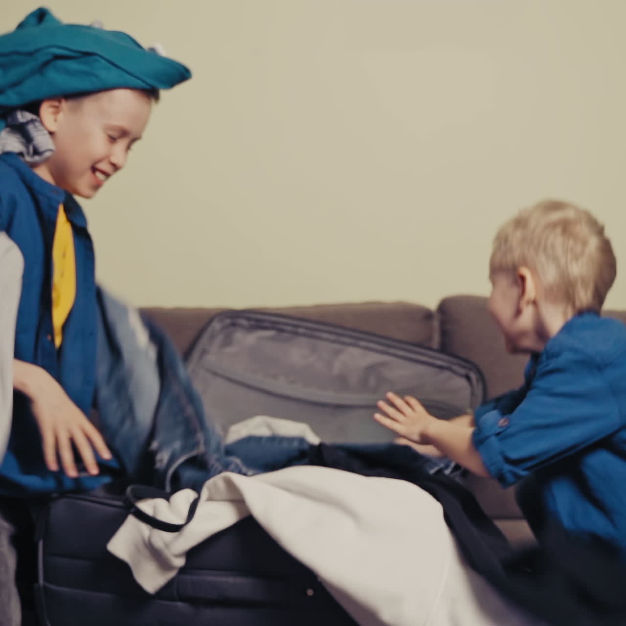 Joyful boys wearing identically are taking out clothes of the packed suitcase. Happy brothers making a mess with clothes on the couch in the room. Slow motion.