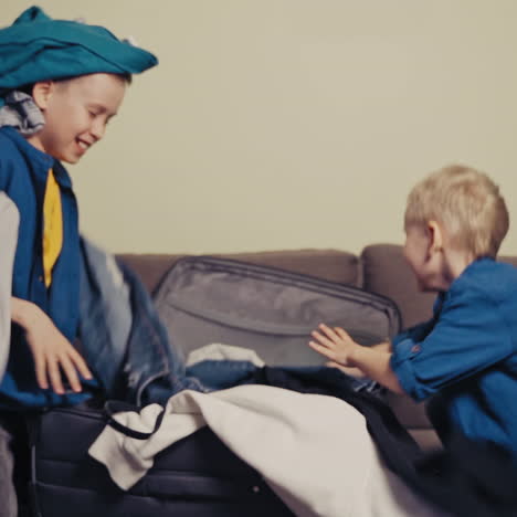 joyful boys wearing identically are taking out clothes of the packed suitcase. happy brothers making a mess with clothes on the couch in the room. slow motion.