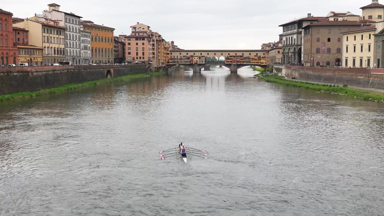 Ampia inquadratura di canottieri che scivolano sull'Arno sotto l'iconico Ponte Vecchio, incorniciati dalla storica architettura di Firenze sotto una luce diurna morbida e un cielo coperto—ideale per riprese di stile di vita urbano o di viaggio.
