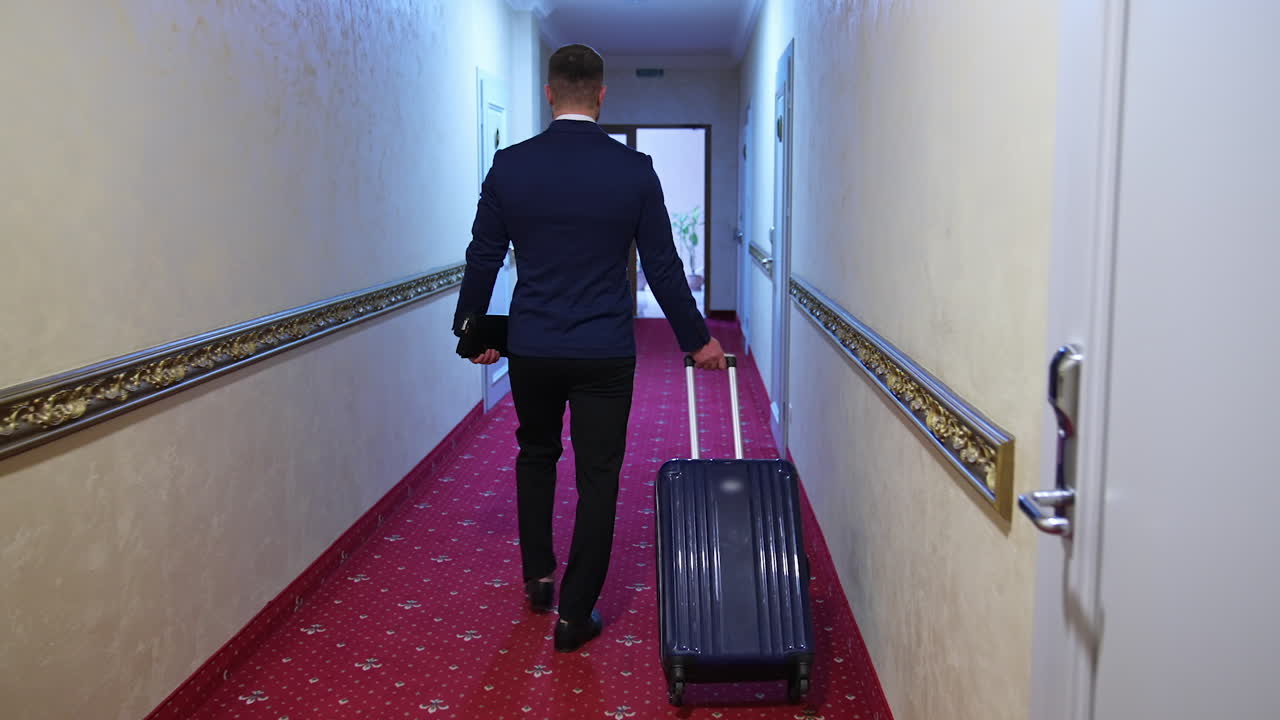 Man in suit with suitcase in hotel. Back view of a businessman with a trolley bag walking to his room in the hotel corridor.