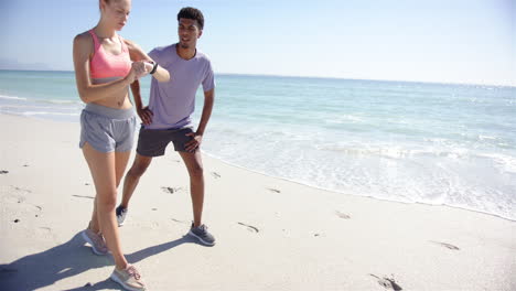 joven mujer caucásica revisa su reloj inteligente al lado de un joven de dos razas en una playa soleada con copia s