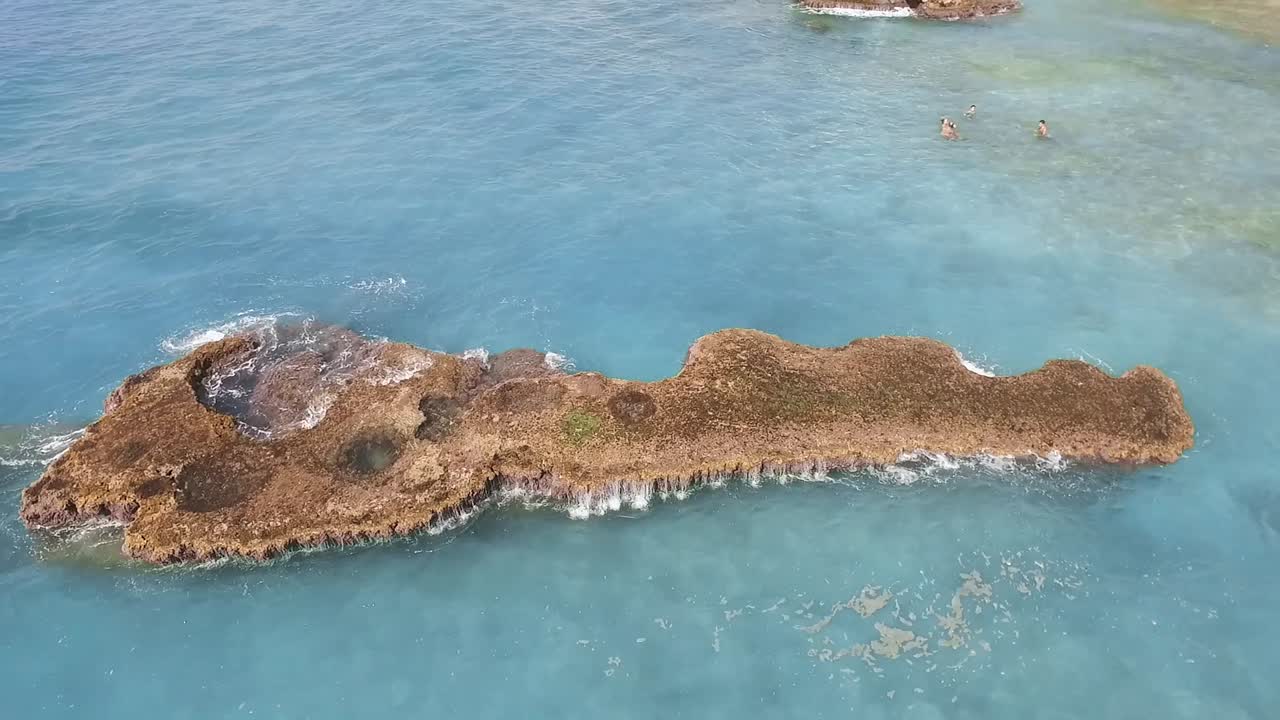 High aerial circle pan rising above long rough rock in blue transparent water near resort