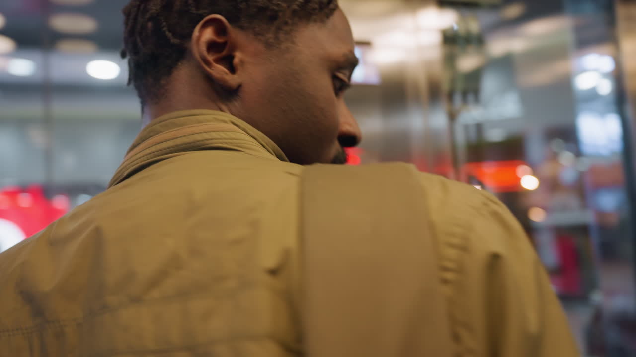 Close up of adult male with braided hairstyle and trimmed beard standing inside elevator, turning head toward glass doors as bright mall lights reflect with people in the mall