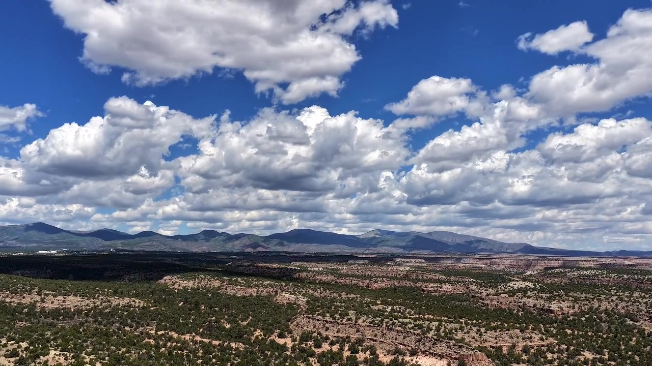 Ampia ripresa aerea con drone in 4K UHD in ascesa che rivela un vasto deserto d'alta quota del Nuovo Messico. Strati di nuvole in rapido movimento attraversano un cielo blu vivido, proiettando ombre dinamiche sulle mesas di roccia rossa a gradoni sottostanti.
