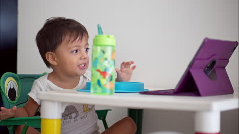 Pequeño-Niño-Latino-Divirtiéndose-Viendo-Un-Espectáculo-En-Una-Tableta-Morada-Mientras-Desayuna-En-Una-Pequeña-Mesa-Blanca-Con-Patas-Coloridas