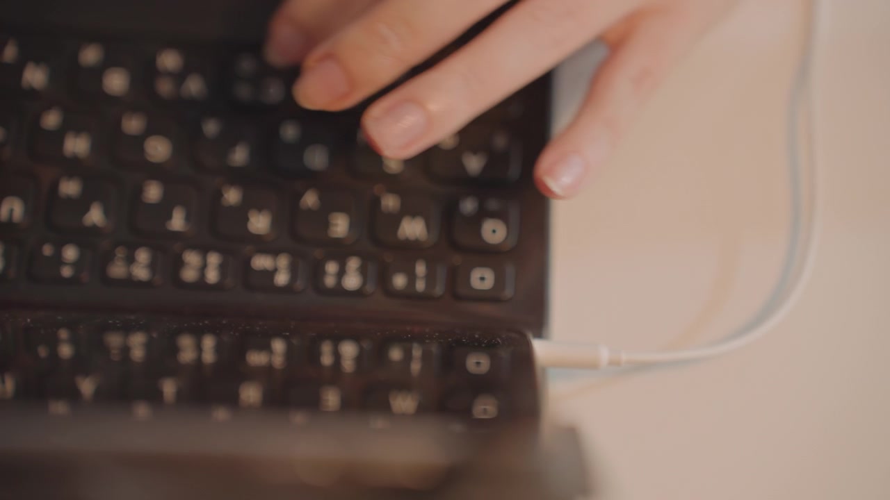 Close up of hands typing on laptop keyboard. Fingers move rapidly over dark keys with white characters reflecting light. Thin cable is visible to right side in later frames
