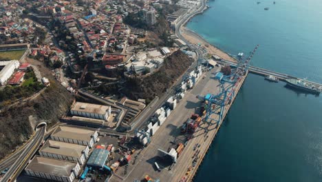 Aerial-orbit-of-containers-and-cranes-in-Valparaiso-Sea-Port,-Cerro-Alegre-hillside-buildings-in-background,-Chile