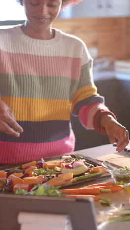 vertical video of happy african american woman preparing meal using tablet in kitchen, slow motion