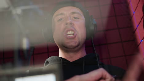 a man dressed in black is wearing headphones and standing in front of a microphone in a red soundproofed room. he is singing with seriousness and moving his hand, conveying deep focus and intensity