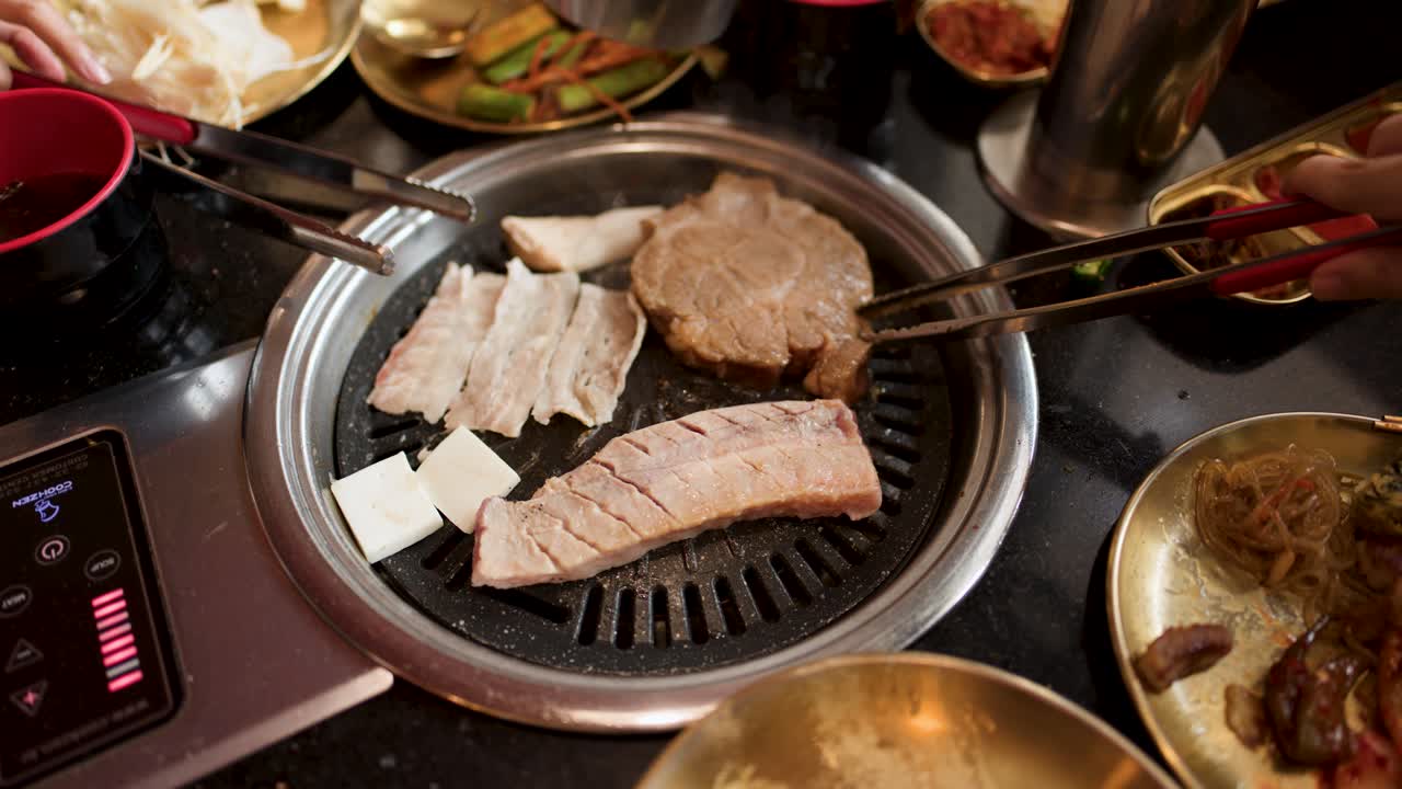 Hand uses tongs to flip pork slices on tabletop Korean barbecue grill under warm lighting