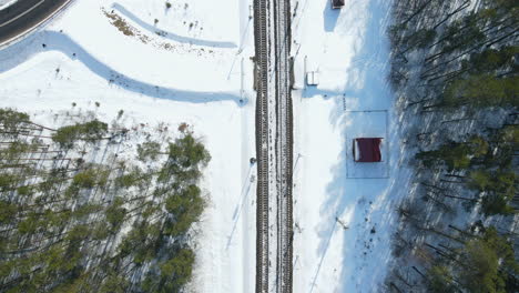 Antena-De-Arriba-Hacia-Abajo-De-Vías-Ferroviarias-Cubiertas-De-Nieve-Vacías,-Pequeña-Estación-De-Tren-Y-Pinos-Durante-El-Día-Soleado-En-Invierno