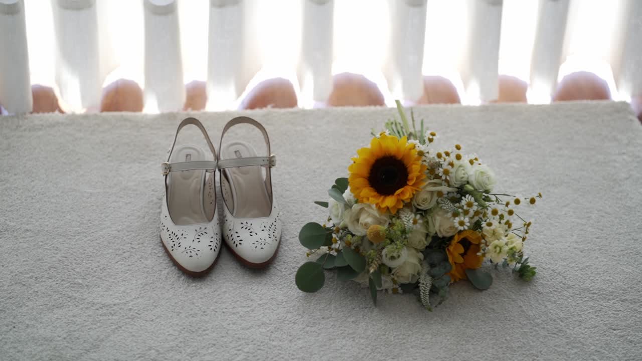 White bridal shoes beside rustic wedding bouquet with sunflower roses and greenery placed on chair in soft window light