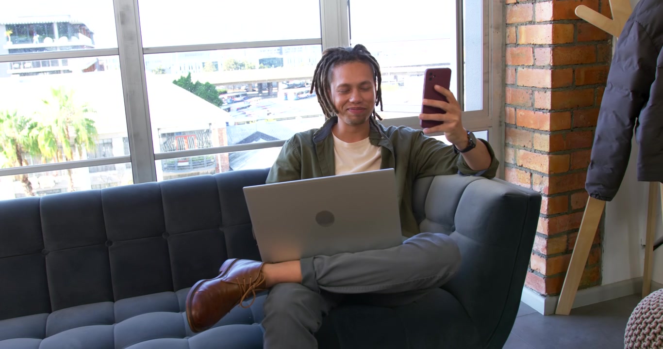 Man receiving call, sitting on tufted-sofa at window, speaking on red phone while working on laptop