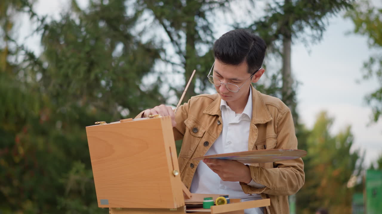 Pelukis Asia di luar ruangan membuat sketsa di atas easel di taman dengan latar belakang pohon pinus, mencampur cat air pada palet, mengenakan jaket cokelat dan kacamata, ekspresi fokus, kedalaman dangkal, suasana hidup tenang, jalanan