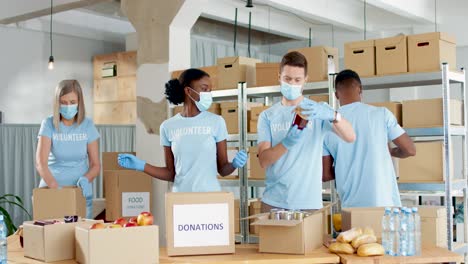 Multiethnic-group-of-volunteers-in-facial-mask-packing-boxes-with-food-in-charity-warehouse