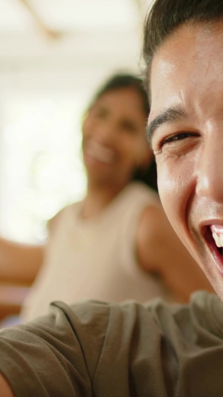 Vertical video: Waving woman in beige top and necklace greeting offscreen, making man laugh at home