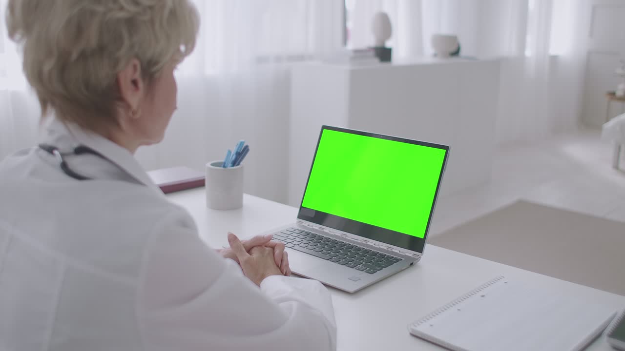 médica de edad está trabajando remotamente, consulta en línea, mirando la pantalla verde de la computadora portátil para la tecnología de clave de croma, telemedicina