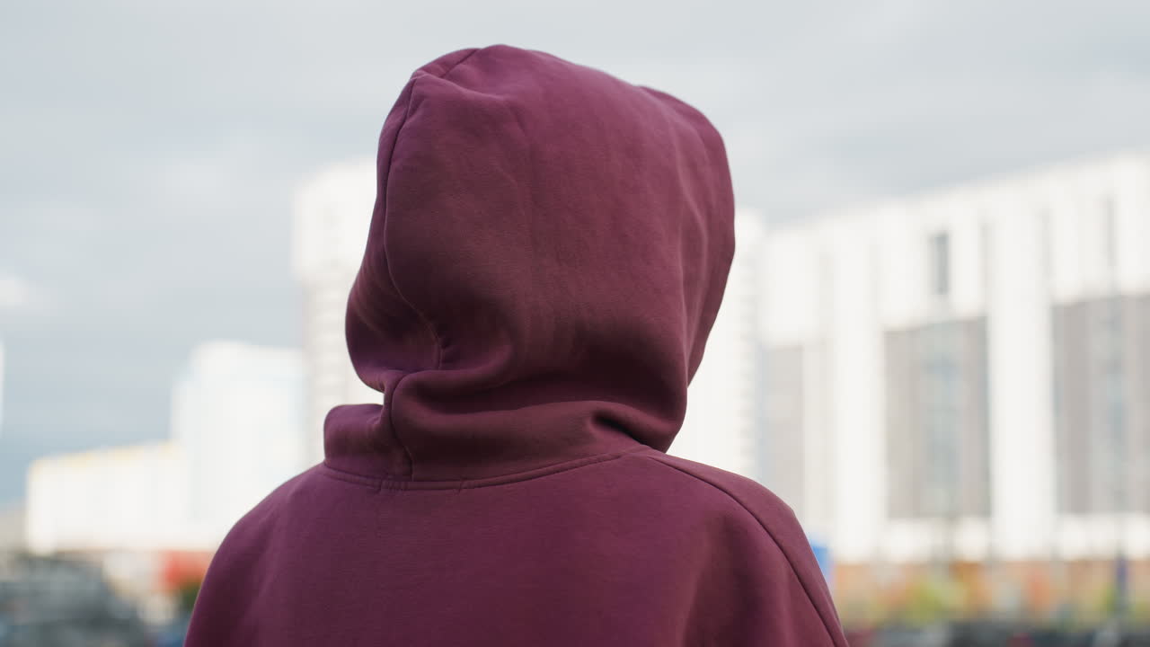 Back view city dweller in wine hoodie with covered hair making fluid motion on urban pavement in front of modern building with subtle blur depth capturing dynamic silhouette and fabric texture