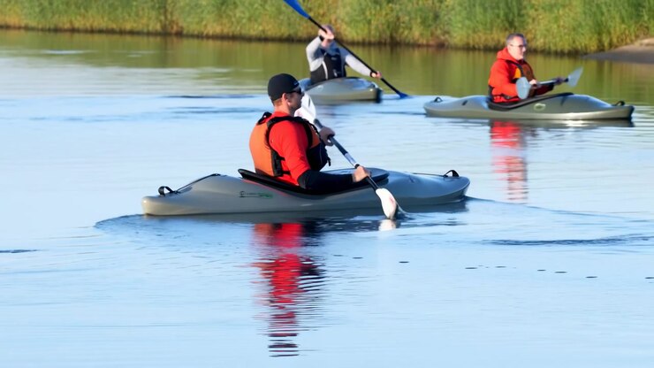 Three kayakers move across calm lake water in staggered diagonal formation while foreground paddler drives forward stroke, rear quarter view shows bright vests, ripples, reflections, and tranquil