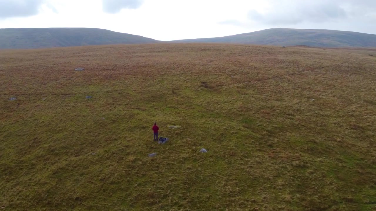 Escursionista in cima a una collina con vista aerea in rapido movimento su un paesaggio di brughiera erbosa sui Brecon Beacons in Galles, Regno Unito, con montagne in lontananza 4K