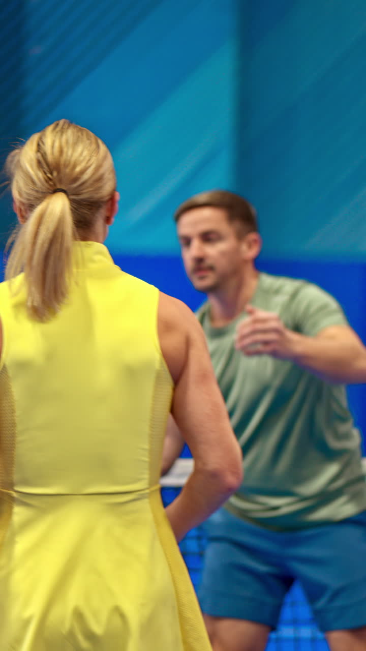 A man and a woman training to play pickleball on a blue, inside court. Vertical