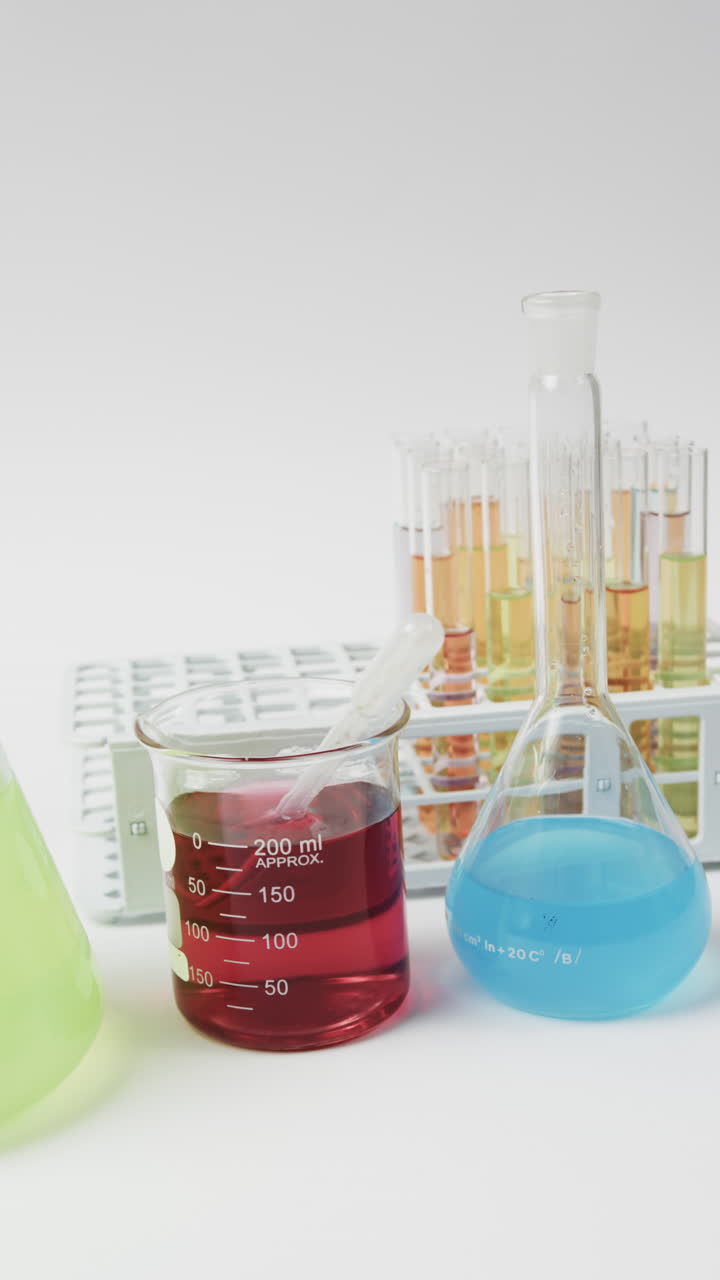 Vertical video of coloured liquids in flasks with test tubes in stand on white background