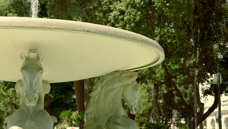 elegant stone horses adorn a large water fountain under the trees on a sunny day