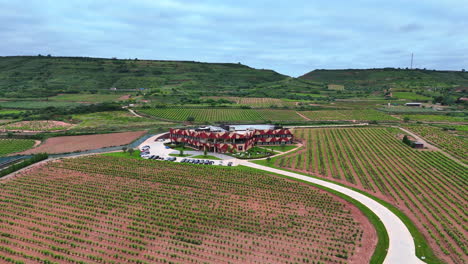 frontal aerial approach to a modern winery building in la rioja, spain, surrounded by lush vineyards and green hills under cloudy sky. concept of wine tourism, viticulture and rural architecture