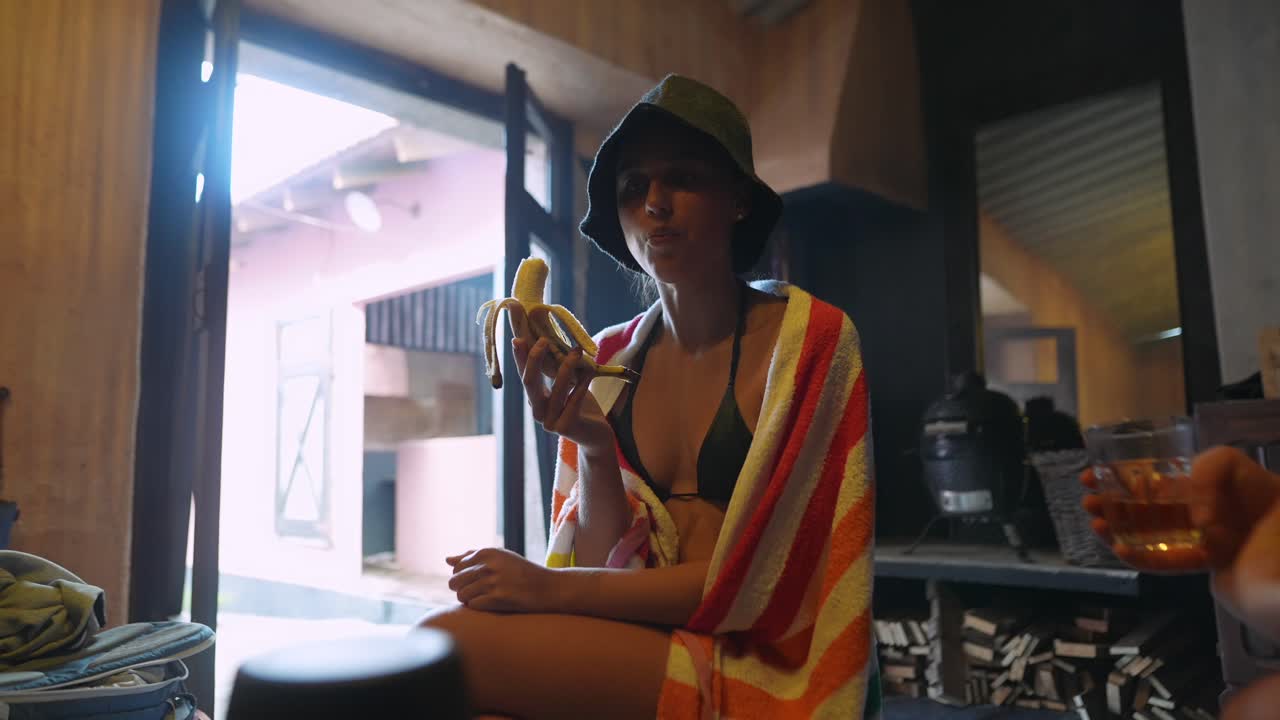 Giovane donna con cappello e asciugamano che mangia una banana al chiuso