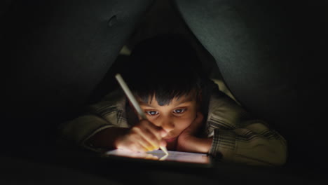 Young-Boy-In-Home-Made-Camp-Made-From-Cushions-Playing-And-Drawing-With-Digital-Tablet-And-Stylus-At-Night-3