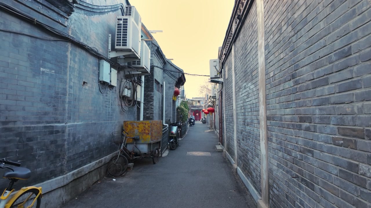 Pekin'in dar hutong sokağında gri tuğla duvarlar, park edilmiş scooterlar ve klima üniteleri bulunuyor. Statik geniş açılı çekim, geleneksel konut sokağını alacakaranlıkta yakalıyor.