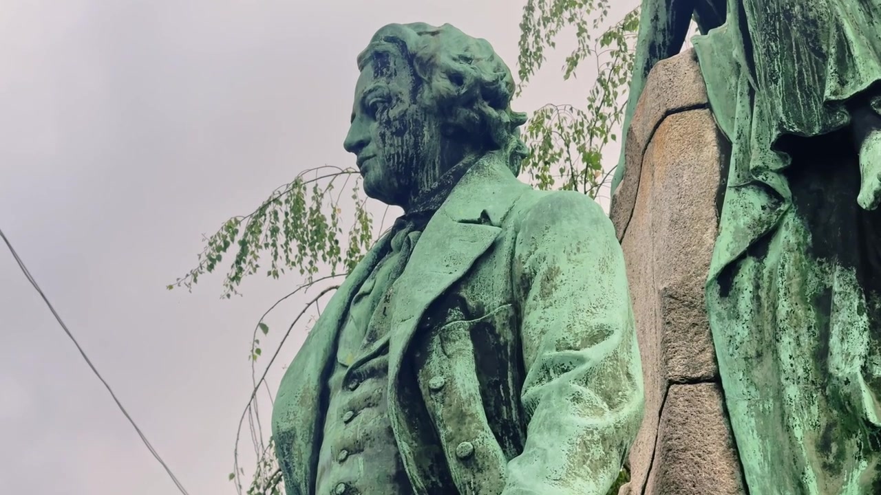 Detailní pohled z nízkého úhlu na sochu France Prešerena v Lublani, zdůrazňující jeho formální kabát, knihu a zvětralý zelený bronzový povrch pod zataženou oblohou.