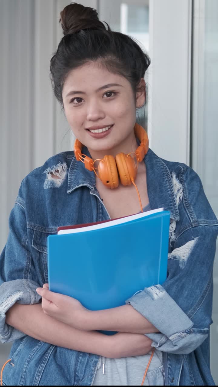 Portrait of an Asian college student holding folders and looking at the camera while standing at the hallway. Education concept.