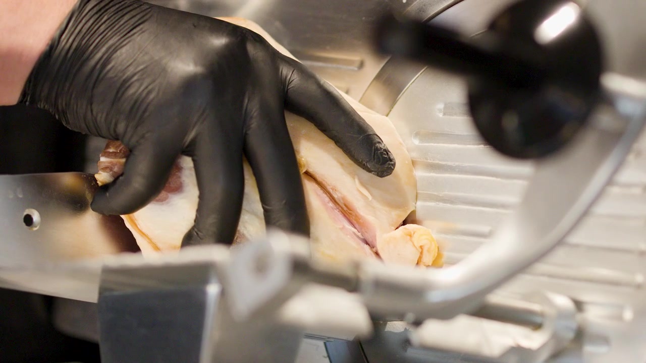 Close-up of gloved hand operating industrial slicer to cut thin meat slices in kitchen.