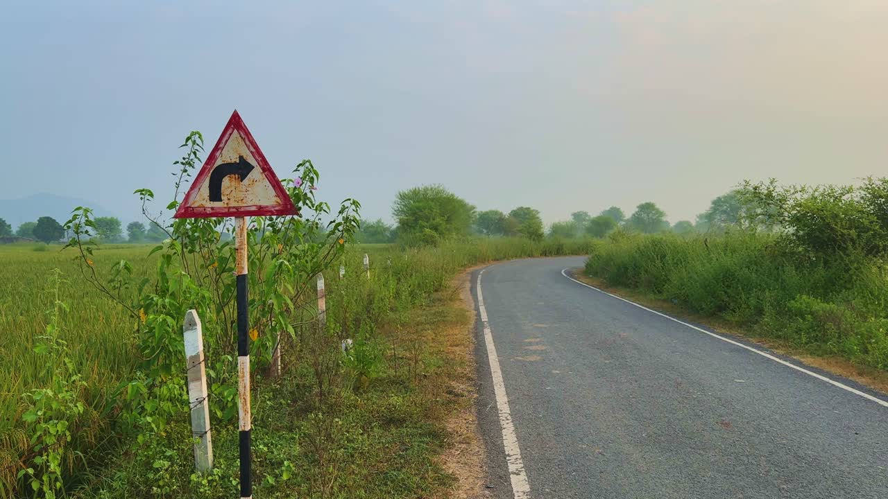 Slow tracking left to right past an old triangular right-turn sign beside a curving rural road bordered by tall green bushes and distant trees under a hazy sky