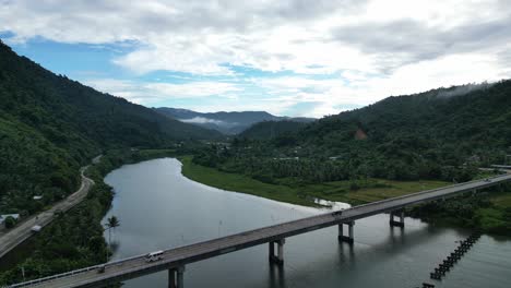 Establishing-Overhead-Drone-Shot-of-Vehicles-driving-across-vast,-idyllic-river-on-long,-modern-bridge-with-jungle-covered-mountains-and-dramatic-clouds-in-background