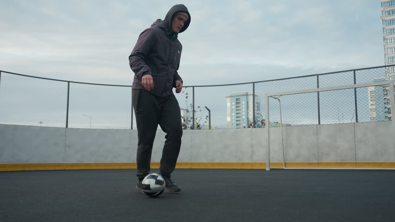 joven controlando hábilmente la pelota de fútbol con un trabajo de pies preciso en la arena deportiva, mostrando equilibrio, agilidad y atletismo, el fondo incluye arquitectura urbana y meta borrosa
