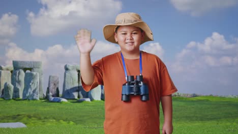 asian boy with a hat and binoculars smiling to camera and waving hand while traveling in stonehenge. boy researcher examines something, travel tourism adventure concept