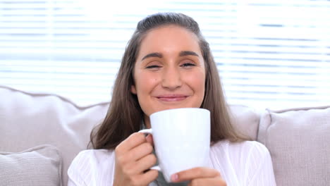 Smiling-brunette-sitting-on-sofa-drinking-her-cup