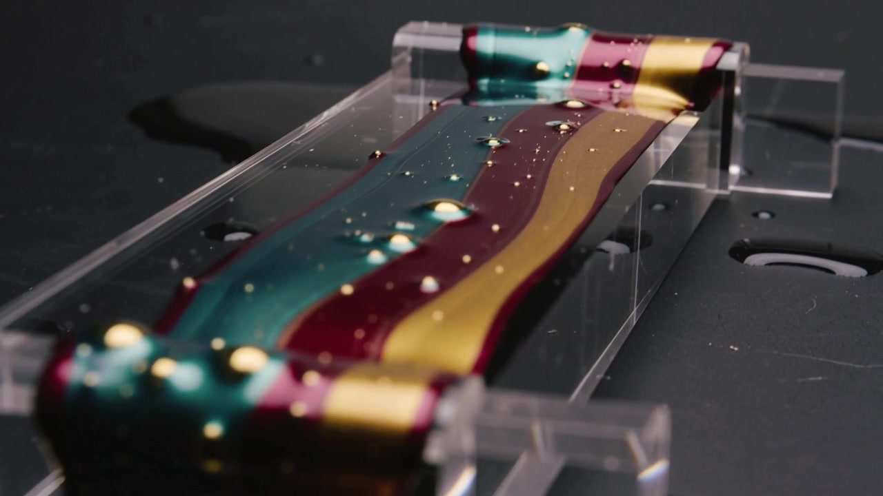 Nastro a strati color verde acqua, magenta e oro che scorre per gravità sul bordo di un canale acrilico e cade su un vassoio nero