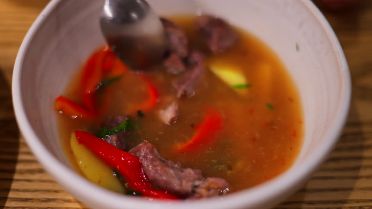 Spoon mixing a broth with chunks of beef, red bell peppers, yellow potatoes, and herbs