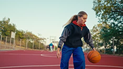 Primer-Plano-De-Una-Chica-Rubia-Con-Uniforme-Deportivo-Que-Corre-Y-Maniobra-Con-Una-Espada-Naranja-Y-Lanza-La-Pelota-Al-Aro-Durante-Su-Juego-De-Baloncesto-En-La-Cancha-De-La-Calle.