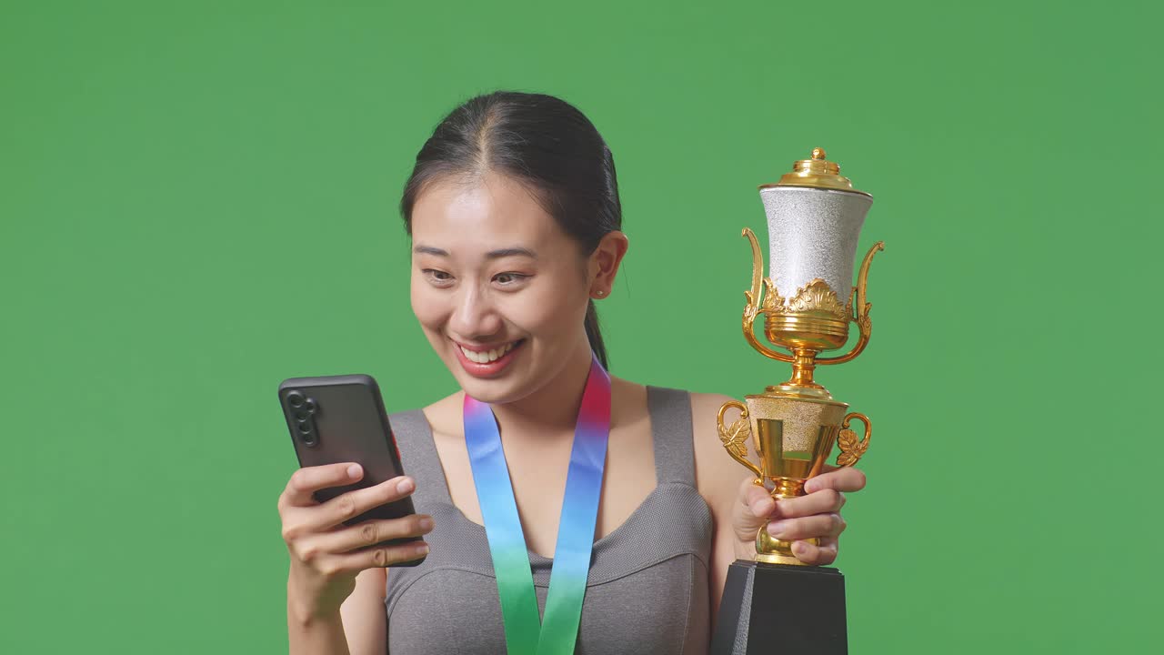 금메달과 트로피를 들고 스마트폰을 보고 있는 아시아 여성의 클로즈업, 그 다음에는 스튜디오의 녹색 스크린 배경에서 축하하는 골을 비명을 지르고 있습니다.