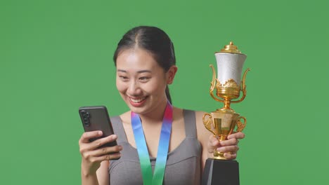 금메달과 트로피를 들고 스마트폰을 보고 있는 아시아 여성의 클로즈업, 그 다음에는 스튜디오의 녹색 스크린 배경에서 축하하는 골을 비명을 지르고 있습니다.