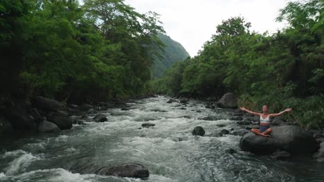 flying through jungle of bali above river with woman on rock in lotus pose, aerial