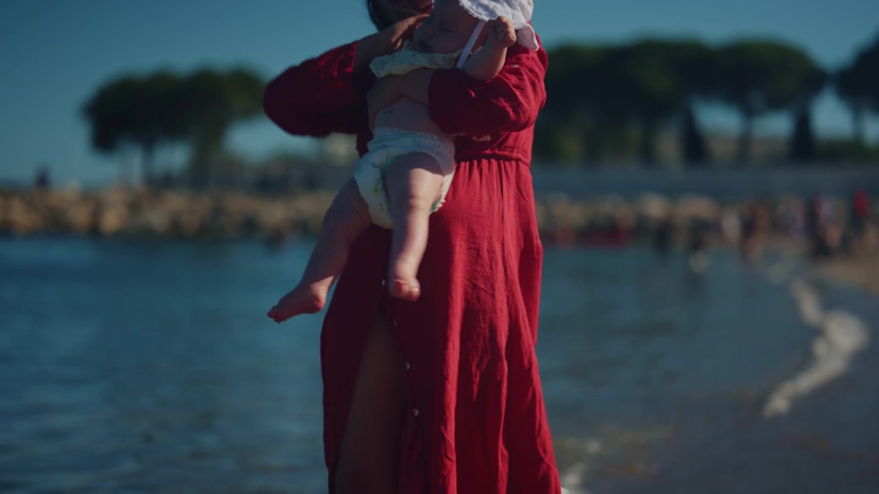 Nærbilde av en mor som støtter babyen sin ved strandkanten mens små bølger skyller over sanden