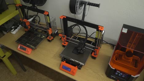 three 3d printers on the table, one of the working on, orange details, black filaments