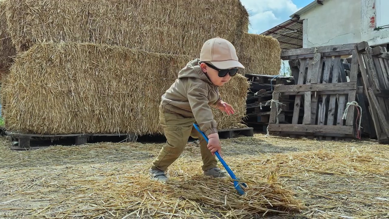 Child plays with hay in a farmyard with bales of hay stacked nearby during a sunny day
