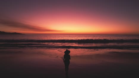 aerial drone 4k flyover woman silhouette walking towards ocean with bright red sunset