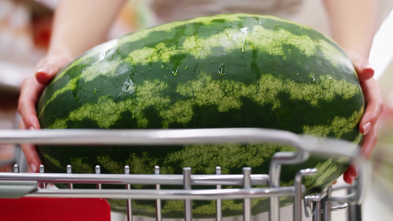 Gros plan montrant des mains stabilisant une pastèque rayée sur les rails d'un chariot métallique, avec un éclairage vif de la zone des fruits et légumes et une composition peu profonde axée sur la texture des fruits frais et l'activité d'achat